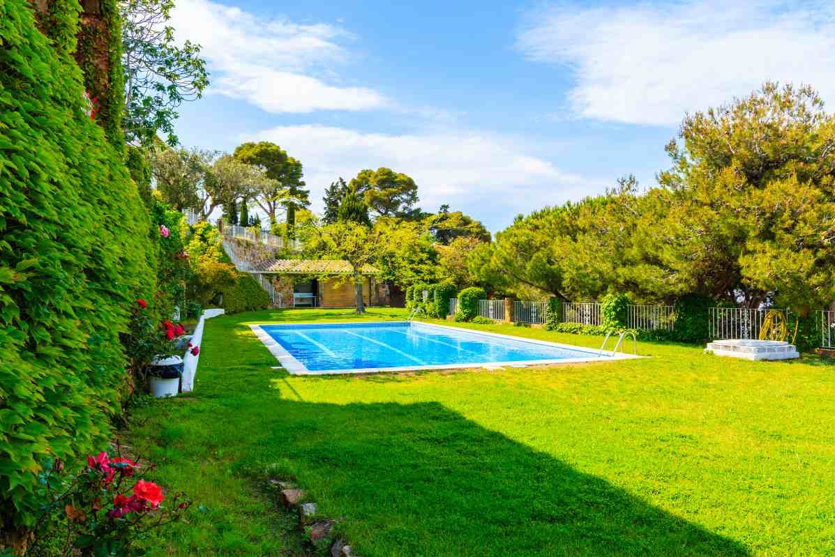 Prostokątny basen z błękitną wodą w ogrodzie z zielonym trawnikiem, otoczony drzewami i roślinnością, z widocznymi schodkami wejściowymi i filtrem piaskowym w tle.