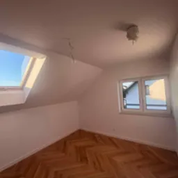Poddasze po remoncie: świeżo pomalowane ściany w bieli, parkiet ułożony w jodełkę, okno dachowe z widokiem na błękitne niebo i okno PCV. Brak opraw oświetleniowych.