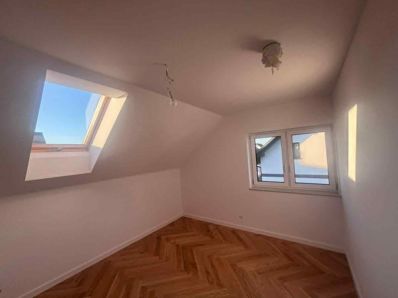 Poddasze po remoncie: świeżo pomalowane ściany w bieli, parkiet ułożony w jodełkę, okno dachowe z widokiem na błękitne niebo i okno PCV. Brak opraw oświetleniowych.