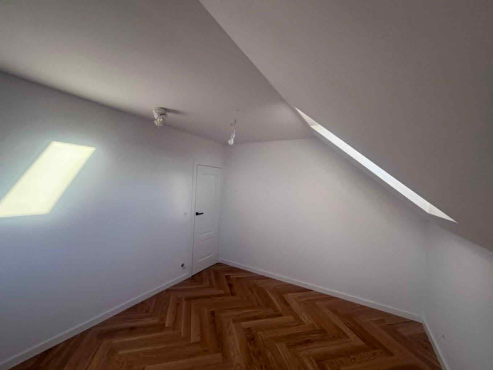 Wnętrze poddasza po remoncie: białe ściany, parkiet w jodełkę, okno dachowe wpuszczające światło, widoczne punkty oświetleniowe i białe drzwi.