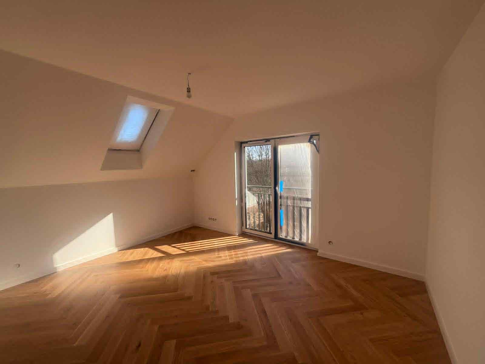 Jasny pokój na poddaszu z parkietem w jodełkę, oknem dachowym i balkonem z widokiem na drzewa. Świeżo pomalowane ściany w neutralnym odcieniu, wpadające promienie słoneczne.