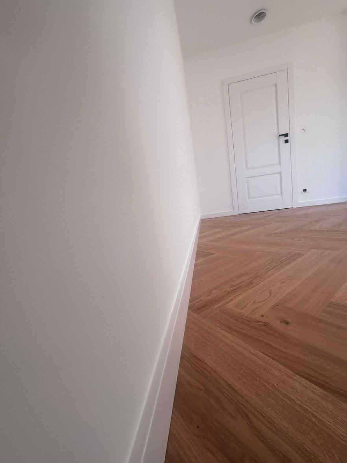 Świeżo pomalowane, białe ściany z widoczną strukturą i ułożona parkietowa podłoga w jodełkę, z białą stolarką drzwiową i listwami przypodłogowymi.