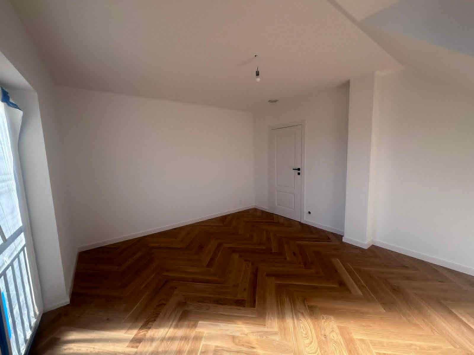 Jasne wnętrze po remoncie: parkiet w jodełkę, świeżo pomalowane na biało ściany i sufit, białe drzwi. Widok z okna z metalową barierką.