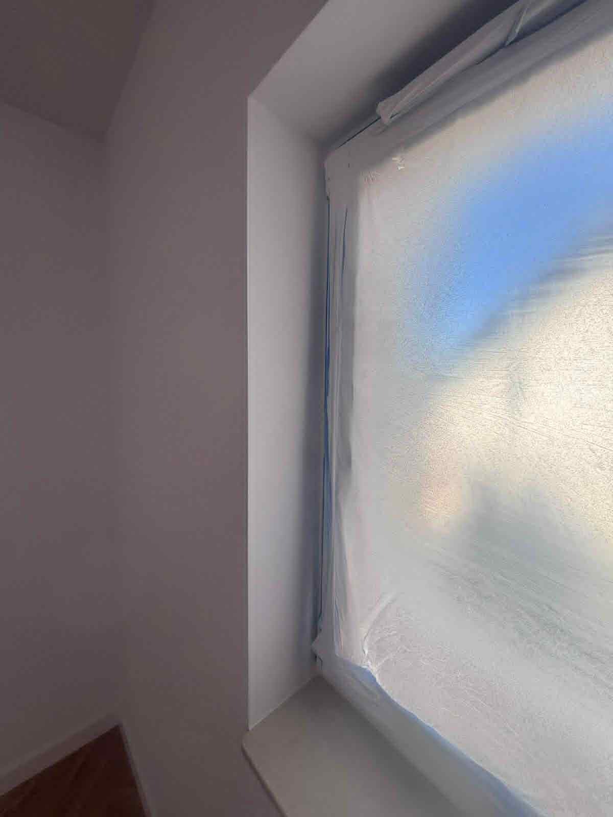 Pokój po malowaniu na biało, okno zabezpieczone folią malarską. Świeżo pomalowana ściana w jasnym odcieniu bieli. Widoczne fragmenty podłogi.