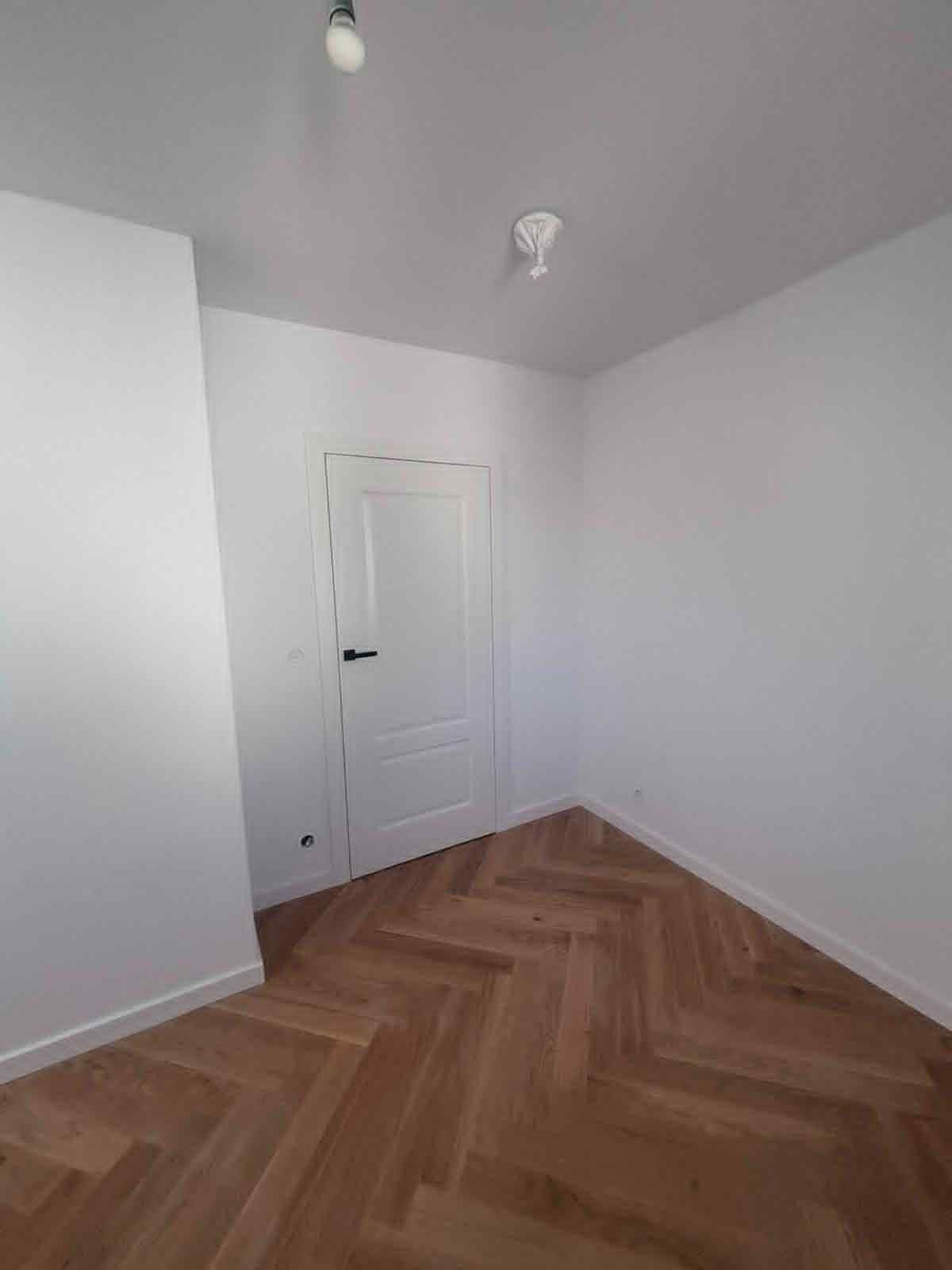 Jasne wnętrze po remoncie: świeżo pomalowane białe ściany, ułożony parkiet w jodełkę, białe drzwi z czarną klamką, widoczne gniazdko elektryczne.