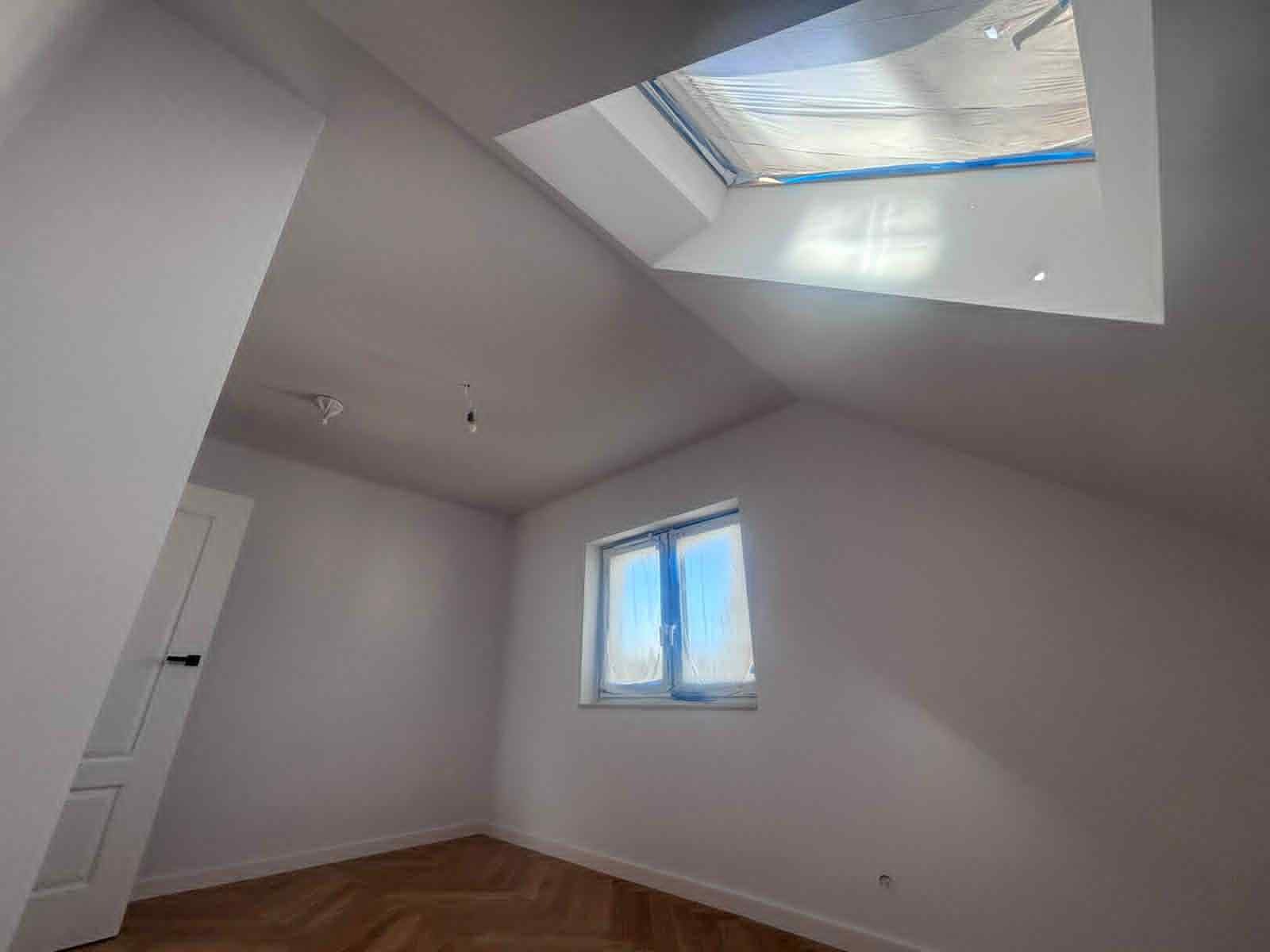 Pomalowane na biało poddasze ze skośnym dachem, oknem połaciowym zabezpieczonym folią, oknem i parkietem. Widoczny fragment drzwi. Świeżo po malowaniu.