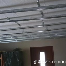 Zysk Remonty - Montaż metalowej konstrukcji sufitu podwieszanego z płyt kartonowo-gipsowych, widoczny szkielet z profili na tle białego sufitu i fragmentu ściany.