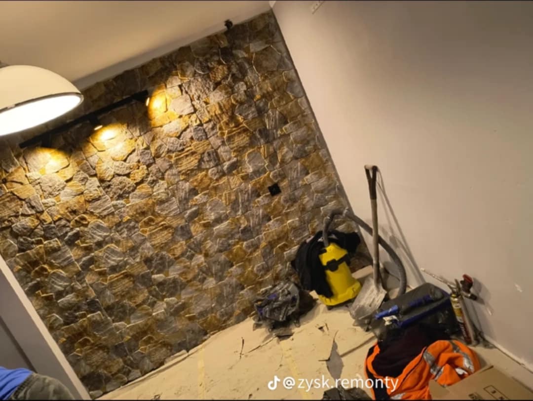 Ściana wykończona dekoracyjnym kamieniem elewacyjnym w odcieniach szarości i brązu, oświetlona reflektorami, z widocznymi narzędziami i odkurzaczem na podłodze w trakcie prac wykończeniowych.