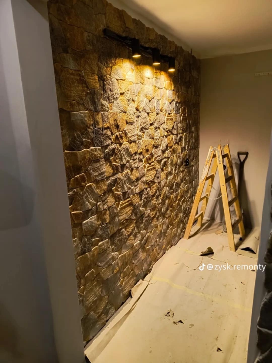 Ściana wykończona kamieniem dekoracyjnym w odcieniach brązu i beżu, oświetlona czarną listwą z reflektorami, widoczna drabina i zabezpieczona podłoga.