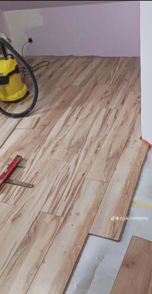 Układanie paneli podłogowych z widocznym poziomem i odkurzaczem na tle ściany w odcieniach fioletu.