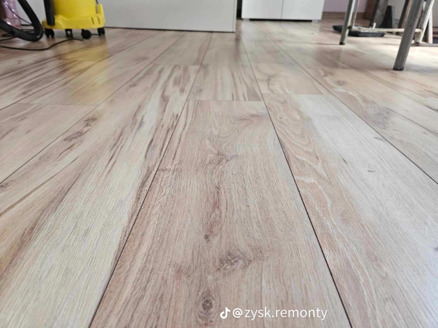 Jasne panele podłogowe ułożone wzdłużnie, widoczne z niskiej perspektywy z odkurzaczem w tle, Klepary.