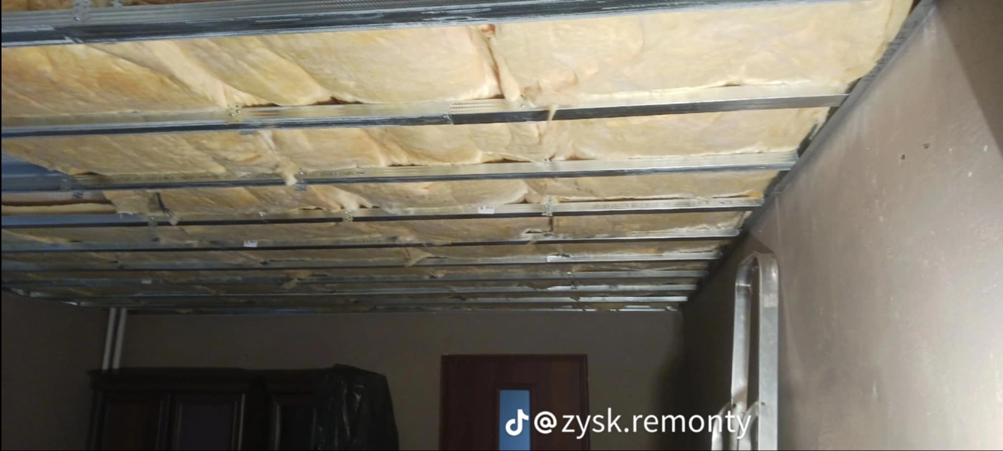 Montaż sufitu podwieszanego z widoczną izolacją z wełny mineralnej, metalowa konstrukcja profili, wnętrze w trakcie prac wykończeniowych.