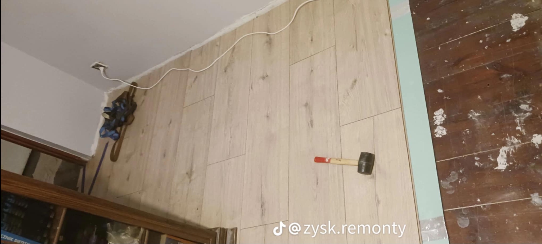 Montaż paneli podłogowych w trakcie, widoczny gumowy młotek, listwa przypodłogowa i fragment starej, zabrudzonej podłogi.