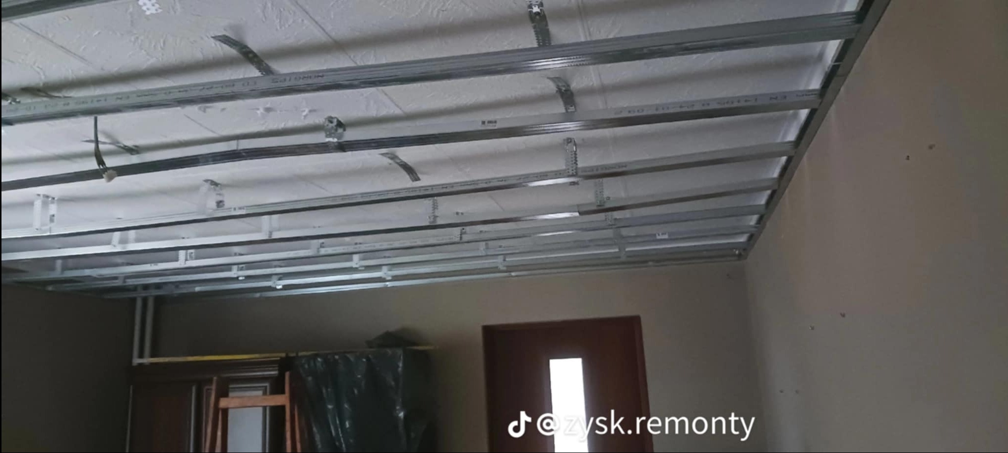 Montaż metalowej konstrukcji sufitu podwieszanego z płyt kartonowo-gipsowych, widoczny szkielet z profili na tle białego sufitu i fragmentu ściany.