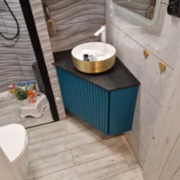 Nowoczesna łazienka z umywalką w kolorze złota na szafce z niebieskimi lamelami, szare płytki z falistym wzorem, drewniane wieszaki w kształcie serc, lustro z oświetleniem i akcesoriami.