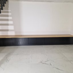 Łukasz Stolarz - Długa, niska szafka z czarnymi ryflowanymi frontami i blatem z jasnego drewna, umieszczona pod schodami z metalową konstrukcją, na podłodze z płytek imitujących marmur.