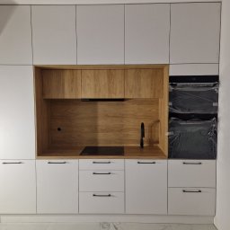 Łukasz Stolarz - Nowoczesna kuchnia z białymi szafkami i drewnianym panelem, czarna armatura i płyta indukcyjna, piekarnik jeszcze w folii ochronnej.