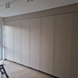 Łukasz Stolarz - Beżowa szafa wnękowa z pionowymi zdobieniami, zamontowana od podłogi do sufitu, widoczna drabinka malarska i parkiet w jodełkę