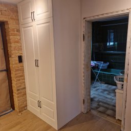 Łukasz Stolarz - Biała, wysoka szafa z frezowanymi drzwiami i czarnymi uchwytami, ustawiona w rogu pomieszczenia z ceglaną ścianą i drewnopodobną podłogą; widok na łazienkę z wanną i suszarką na pranie w tle.