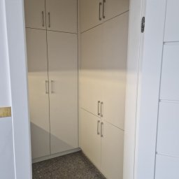 Łukasz Stolarz - Wnętrze garderoby z zabudową meblową w jasnym odcieniu, widok z zewnątrz przez otwarte drzwi, szary lastryko na podłodze.