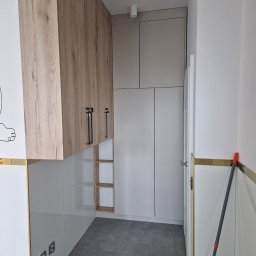 Łukasz Stolarz - Wysoka zabudowa w wąskim korytarzu: białe lakierowane szafy z drewnopodobnymi górnymi szafkami i czarnymi uchwytami, szare płytki podłogowe, listwa przypodłogowa w kolorze złota