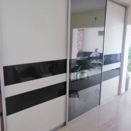 Łukasz Stolarz - Biała szafa przesuwna z czarnymi, poziomymi pasami i lustrzanymi drzwiami, odbijającymi wnętrze salonu z siedzącą osobą.