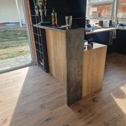 Łukasz Stolarz - Kuchenna wyspa z drewnianym frontem i szarą, betonową ścianką działową, z barkiem i ekspozycją alkoholi, w tle okno z widokiem na budowę.