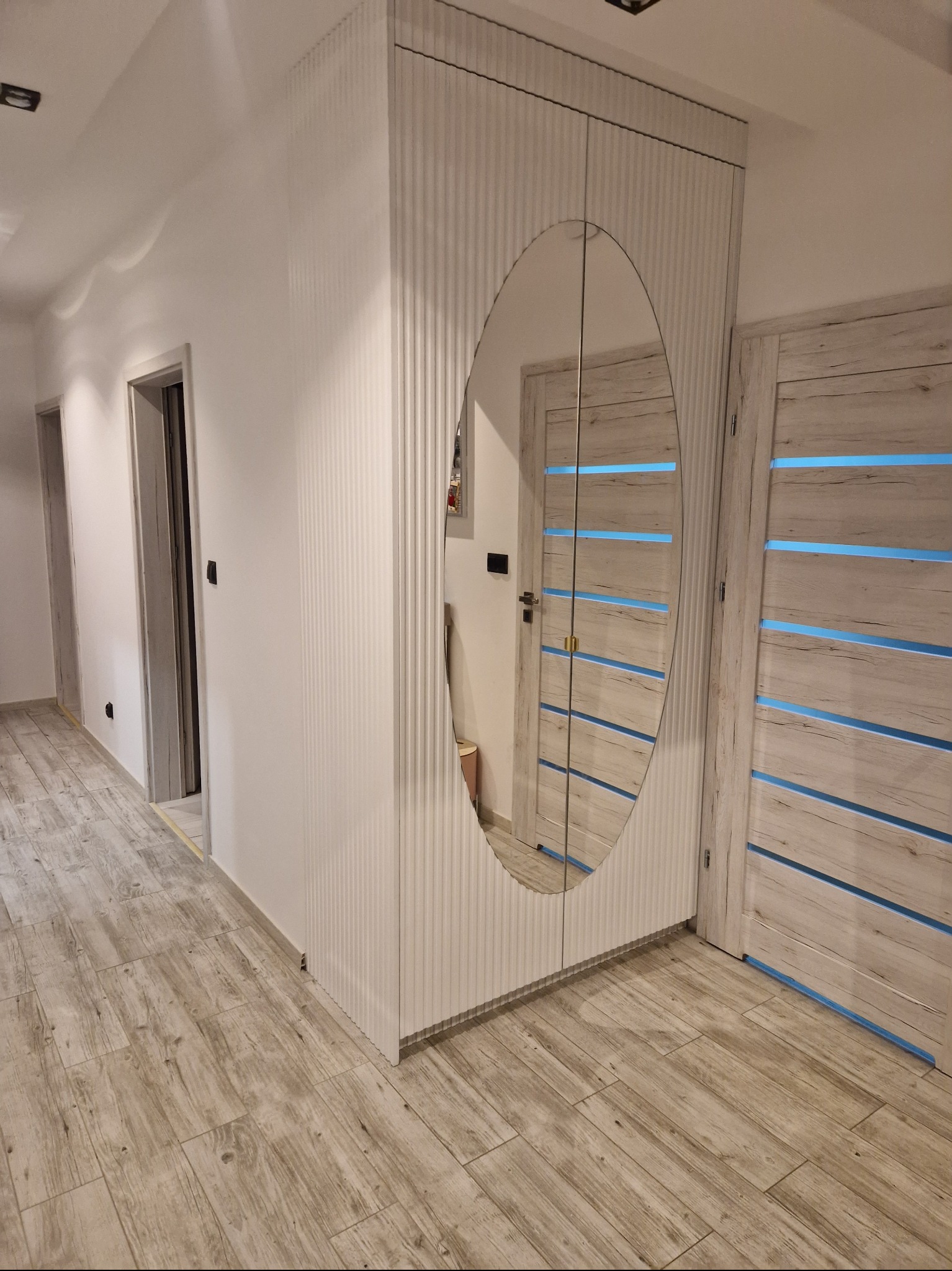 Nowoczesna szafa wnękowa z lustrem w kształcie owalu, białe, ryflowane fronty, drewnopodobna podłoga i drzwi z niebieskimi akcentami w tle.