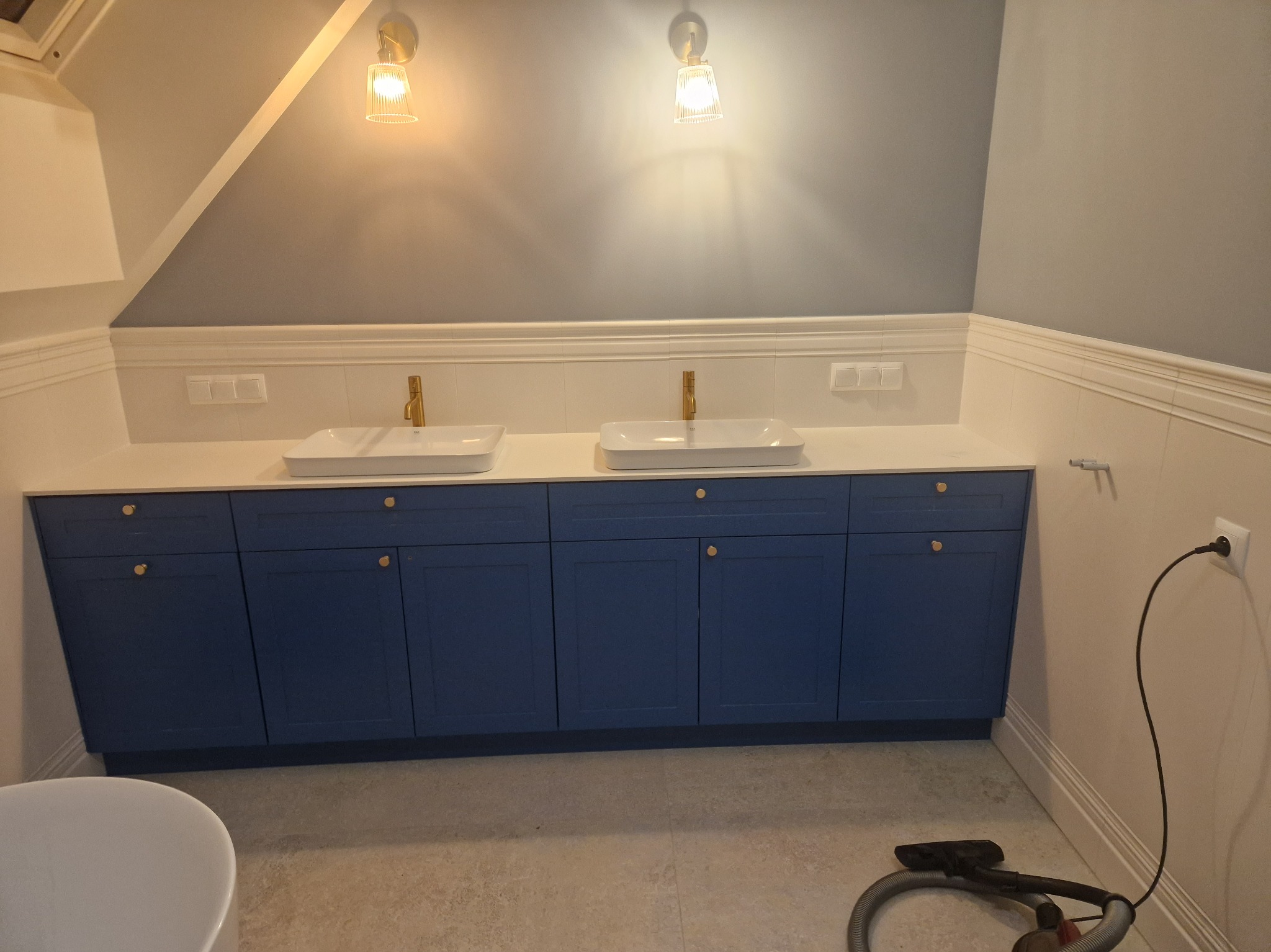 Nowoczesna łazienka na poddaszu z niebieskimi szafkami pod umywalkami, złotymi bateriami i lampami w stylu retro.