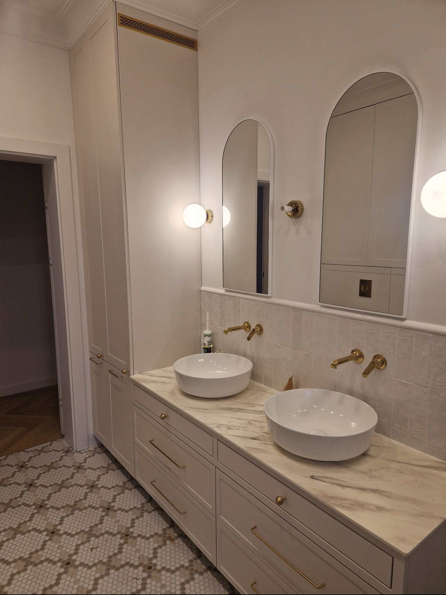 Elegancka łazienka z dwoma umywalkami nablatowymi, marmurowym blatem, złotymi bateriami i okrągłymi lustrami z podświetleniem; wysoka szafka z frontami w kolorze blatu, mozaika na podłodze.