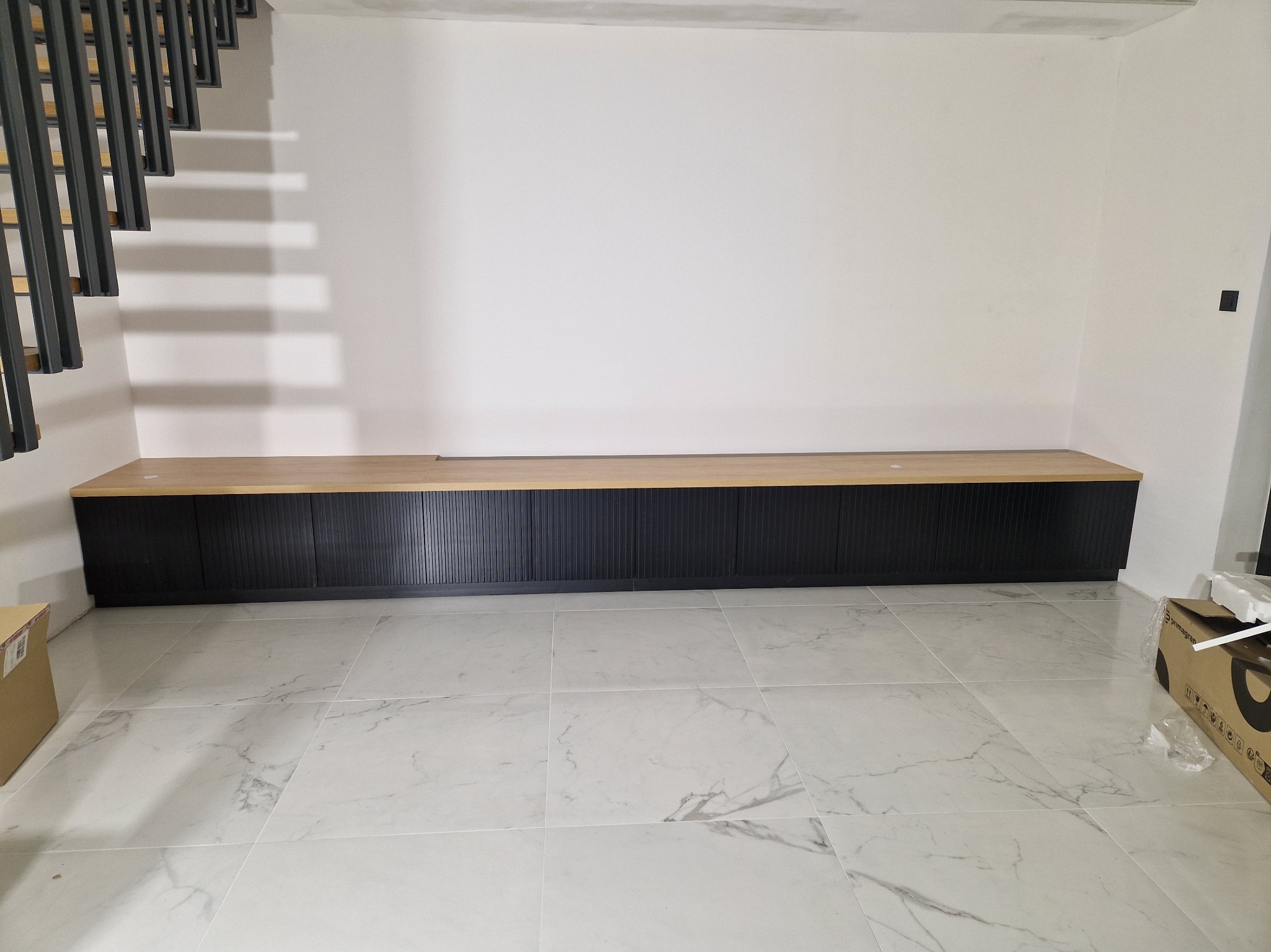 Długa, niska szafka z czarnymi ryflowanymi frontami i blatem z jasnego drewna, umieszczona pod schodami z metalową konstrukcją, na podłodze z płytek imitujących marmur.
