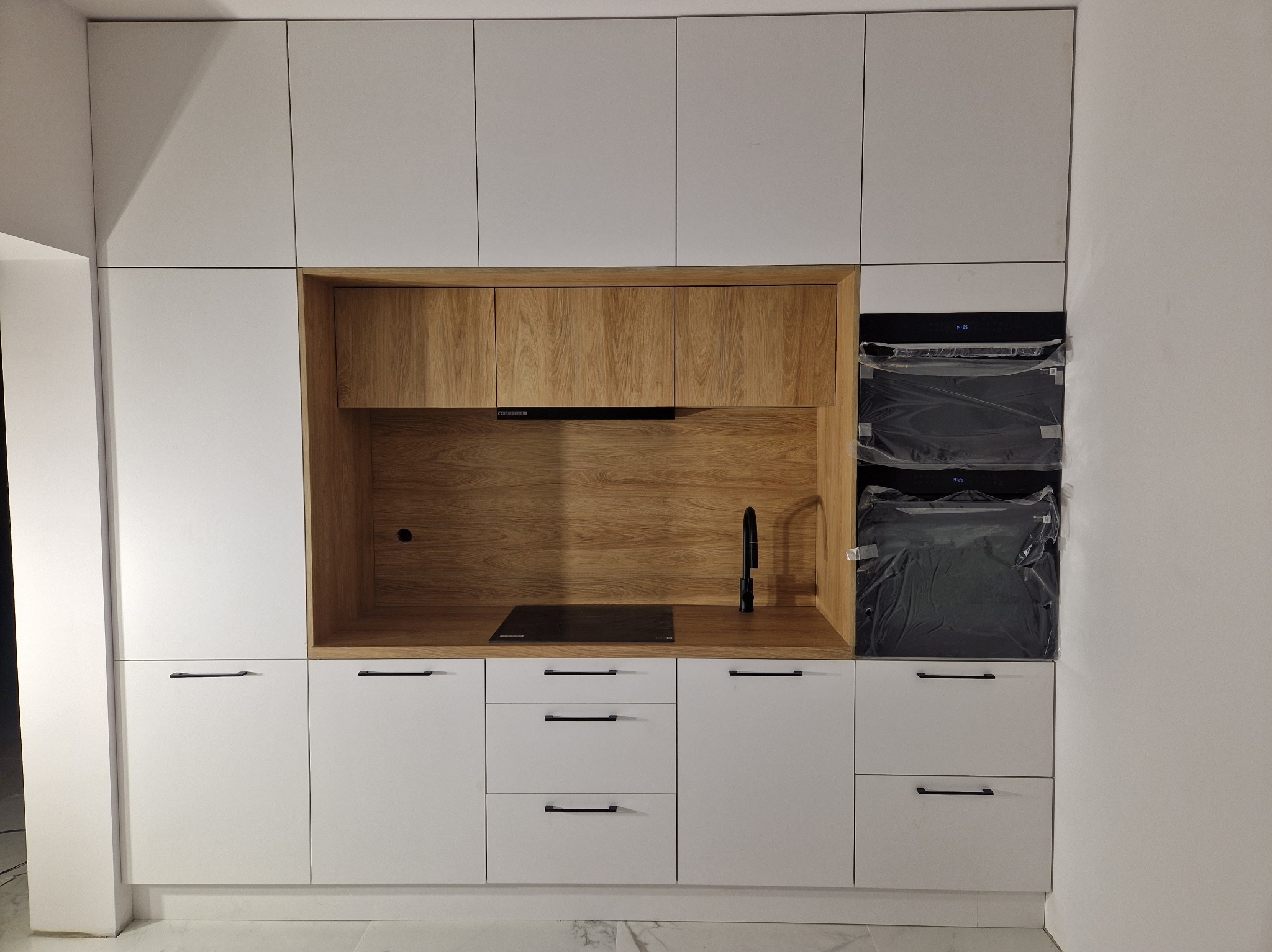 Nowoczesna kuchnia z białymi szafkami i drewnianym panelem, czarna armatura i płyta indukcyjna, piekarnik jeszcze w folii ochronnej.