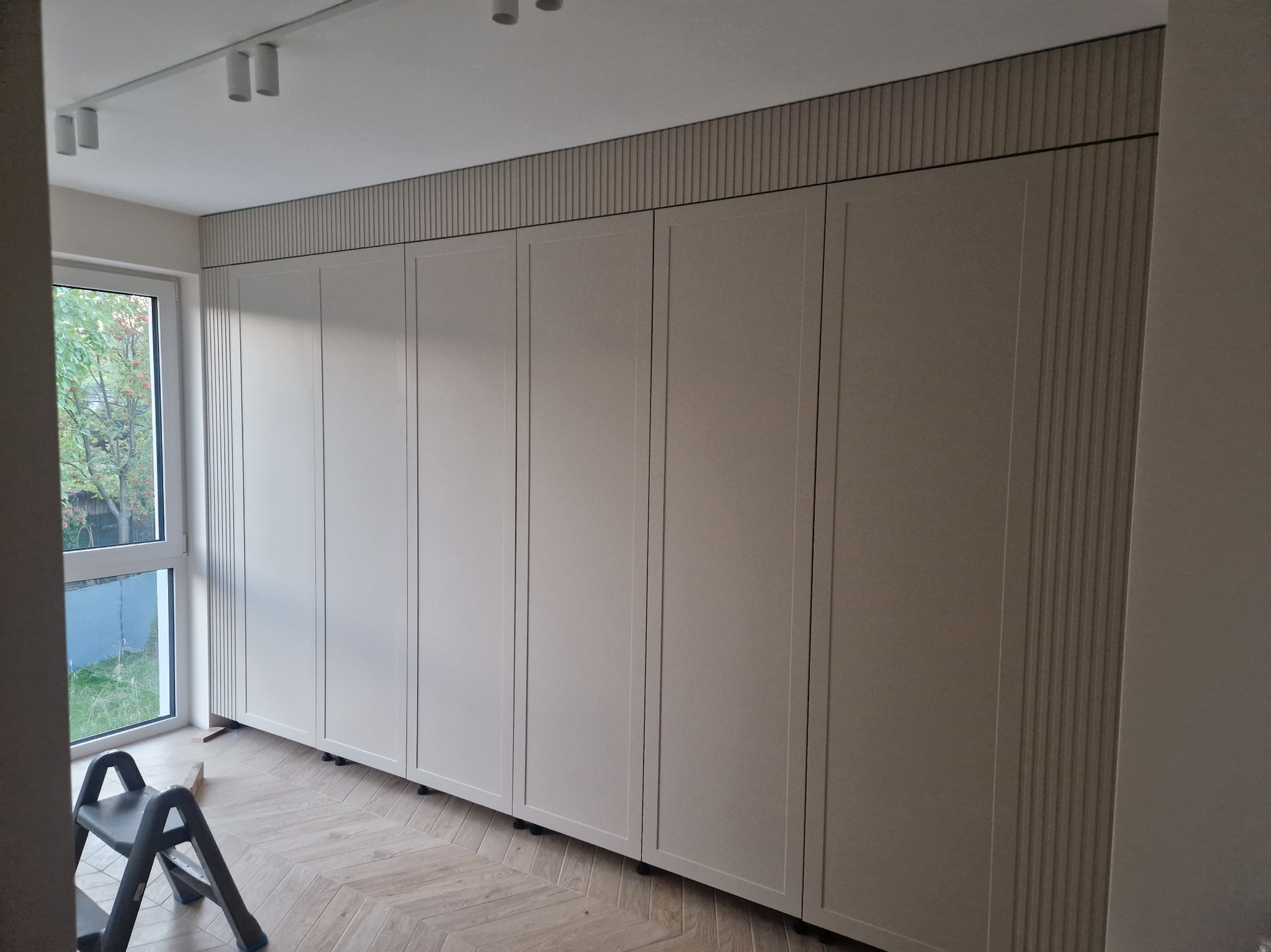 Beżowa szafa wnękowa z pionowymi zdobieniami, zamontowana od podłogi do sufitu, widoczna drabinka malarska i parkiet w jodełkę