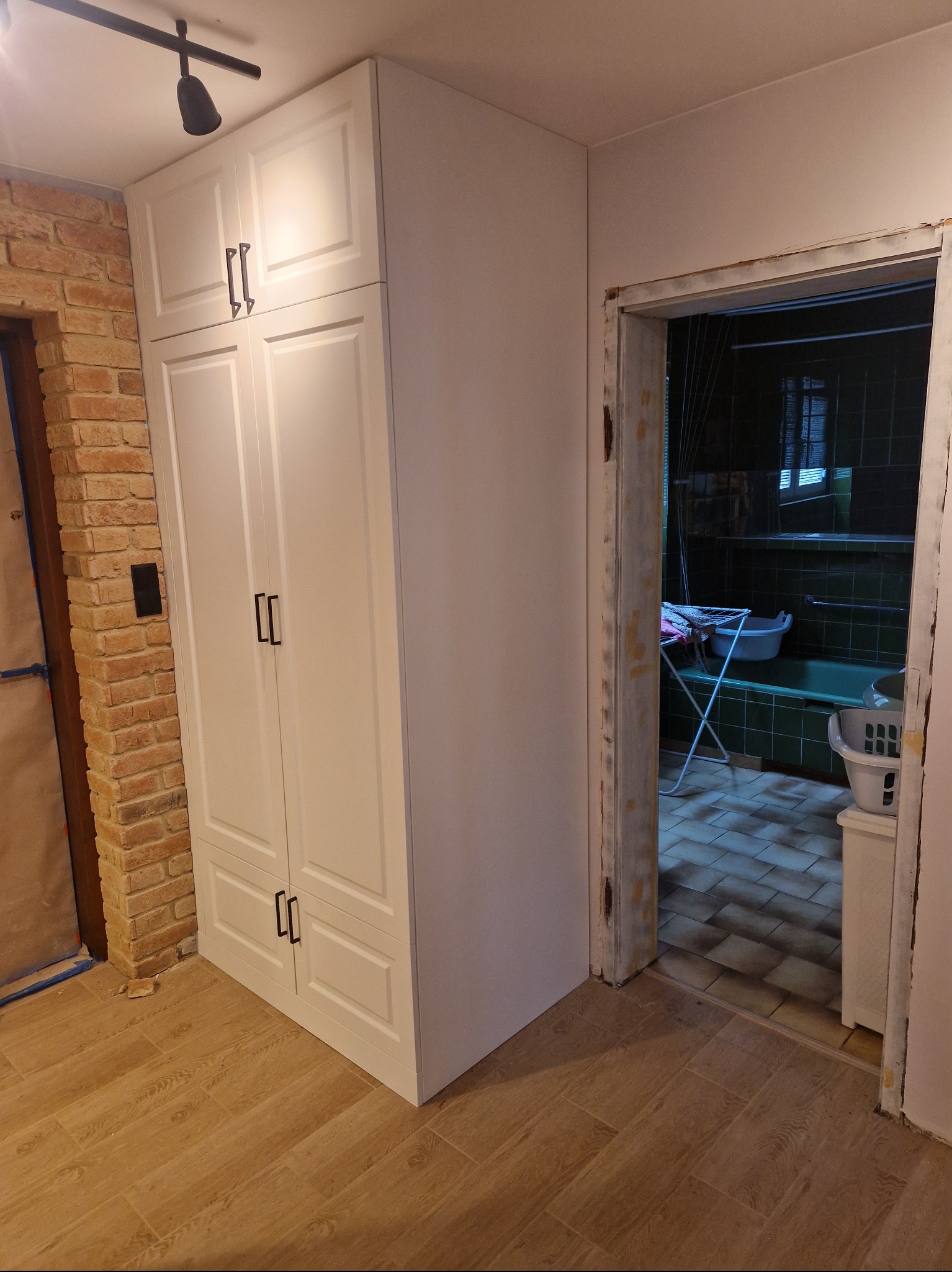 Biała, wysoka szafa z frezowanymi drzwiami i czarnymi uchwytami, ustawiona w rogu pomieszczenia z ceglaną ścianą i drewnopodobną podłogą; widok na łazienkę z wanną i suszarką na pranie w tle.