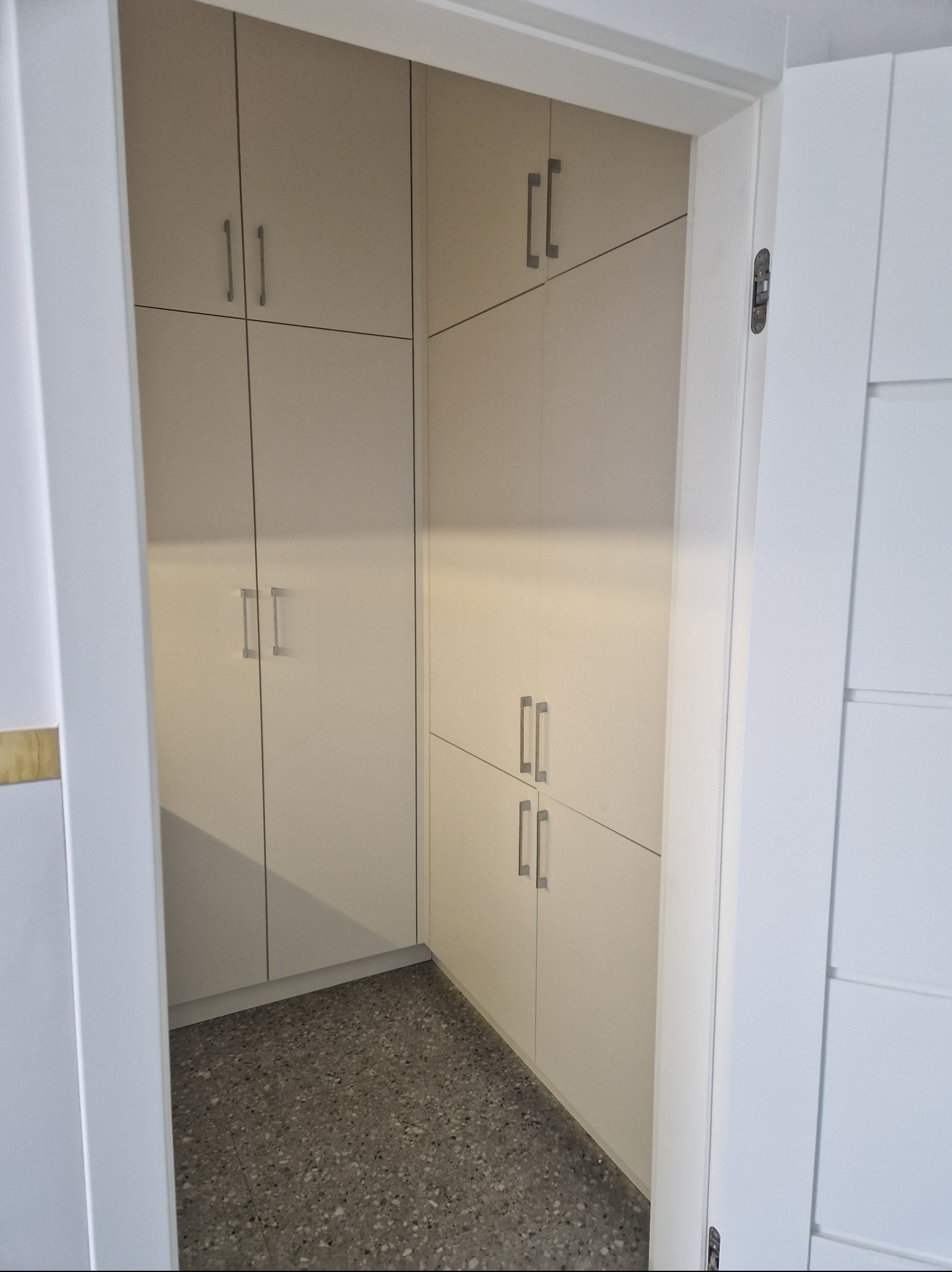 Wnętrze garderoby z zabudową meblową w jasnym odcieniu, widok z zewnątrz przez otwarte drzwi, szary lastryko na podłodze.