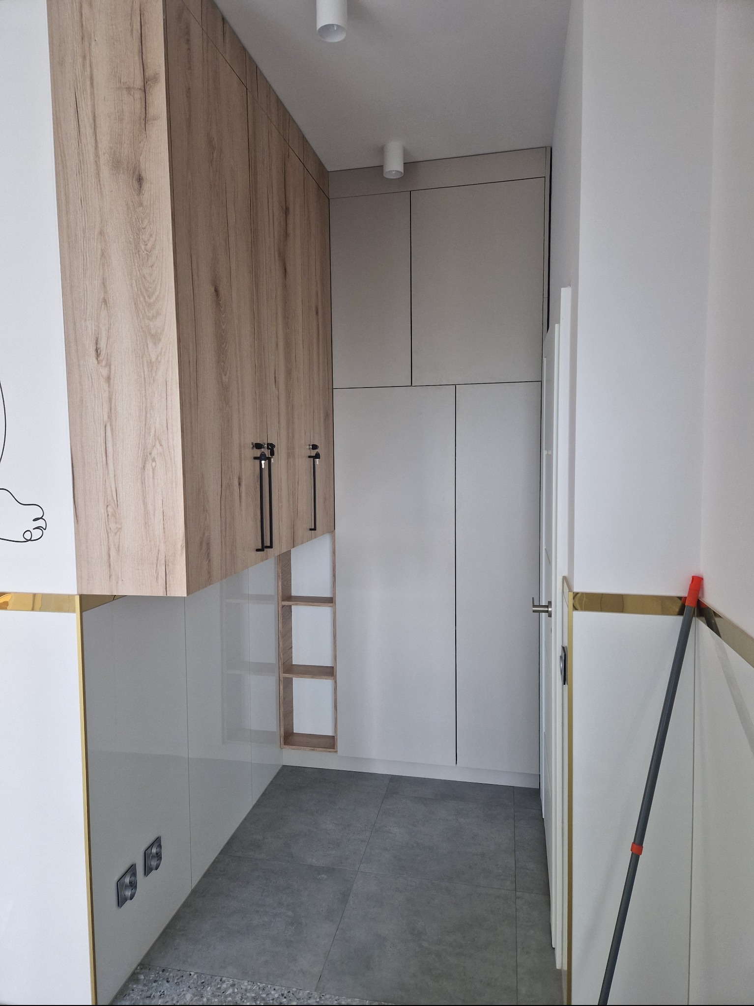 Wysoka zabudowa w wąskim korytarzu: białe lakierowane szafy z drewnopodobnymi górnymi szafkami i czarnymi uchwytami, szare płytki podłogowe, listwa przypodłogowa w kolorze złota