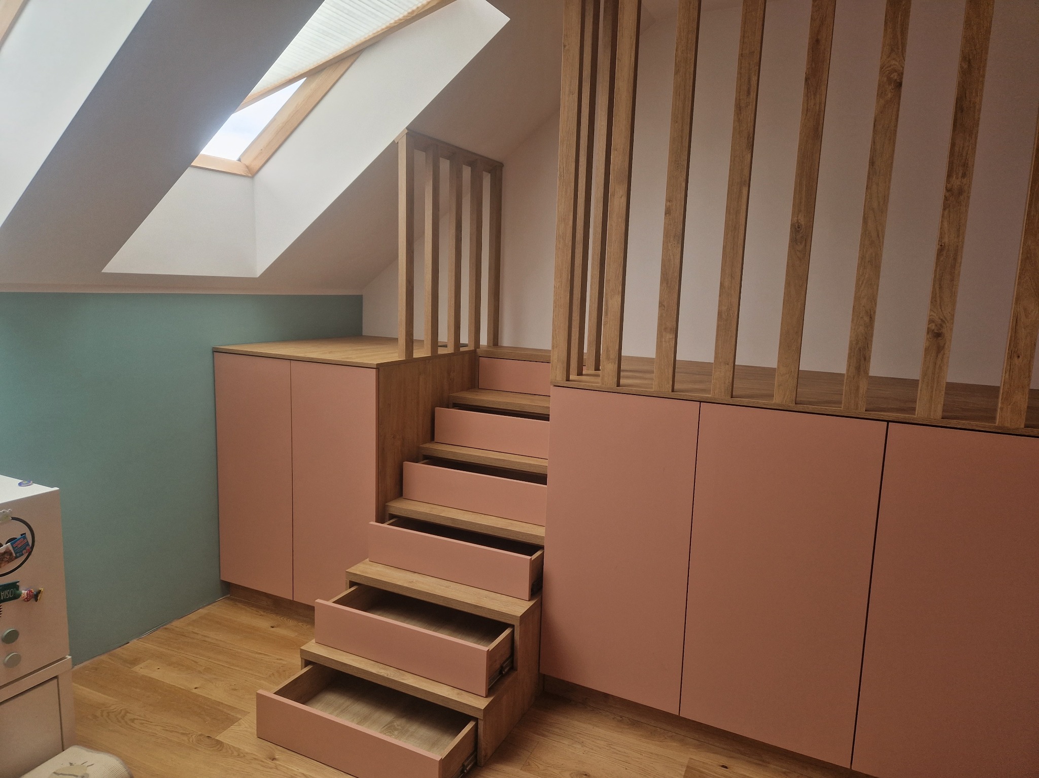 Poddasze z zabudową meblową w odcieniach różu i drewna, schody z szufladami prowadzące na antresolę, pionowe drewniane listwy jako balustrada, okno dachowe.