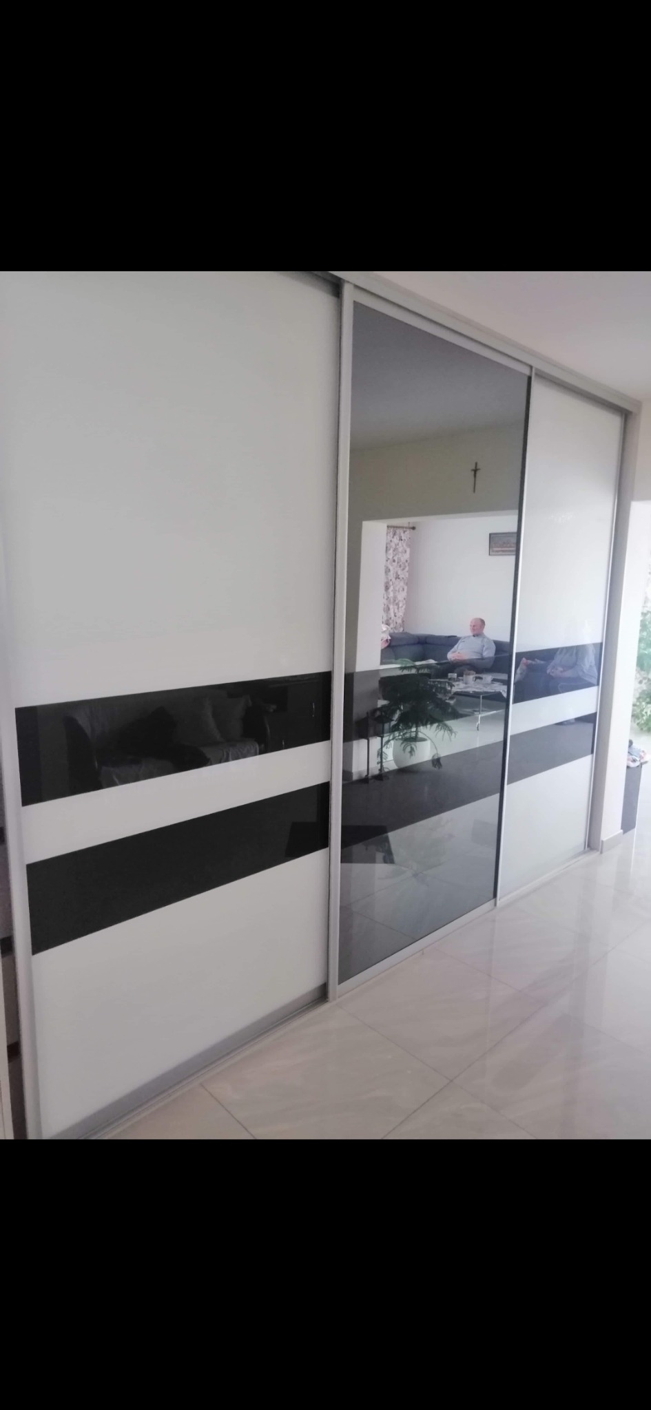 Biała szafa przesuwna z czarnymi, poziomymi pasami i lustrzanymi drzwiami, odbijającymi wnętrze salonu z siedzącą osobą.