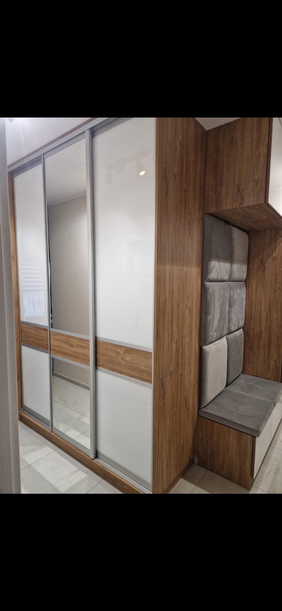 Szafa wnękowa z przesuwnymi drzwiami z lustrem i wbudowanym siedziskiem z tapicerowanym panelem ściennym w odcieniach szarości i brązu.