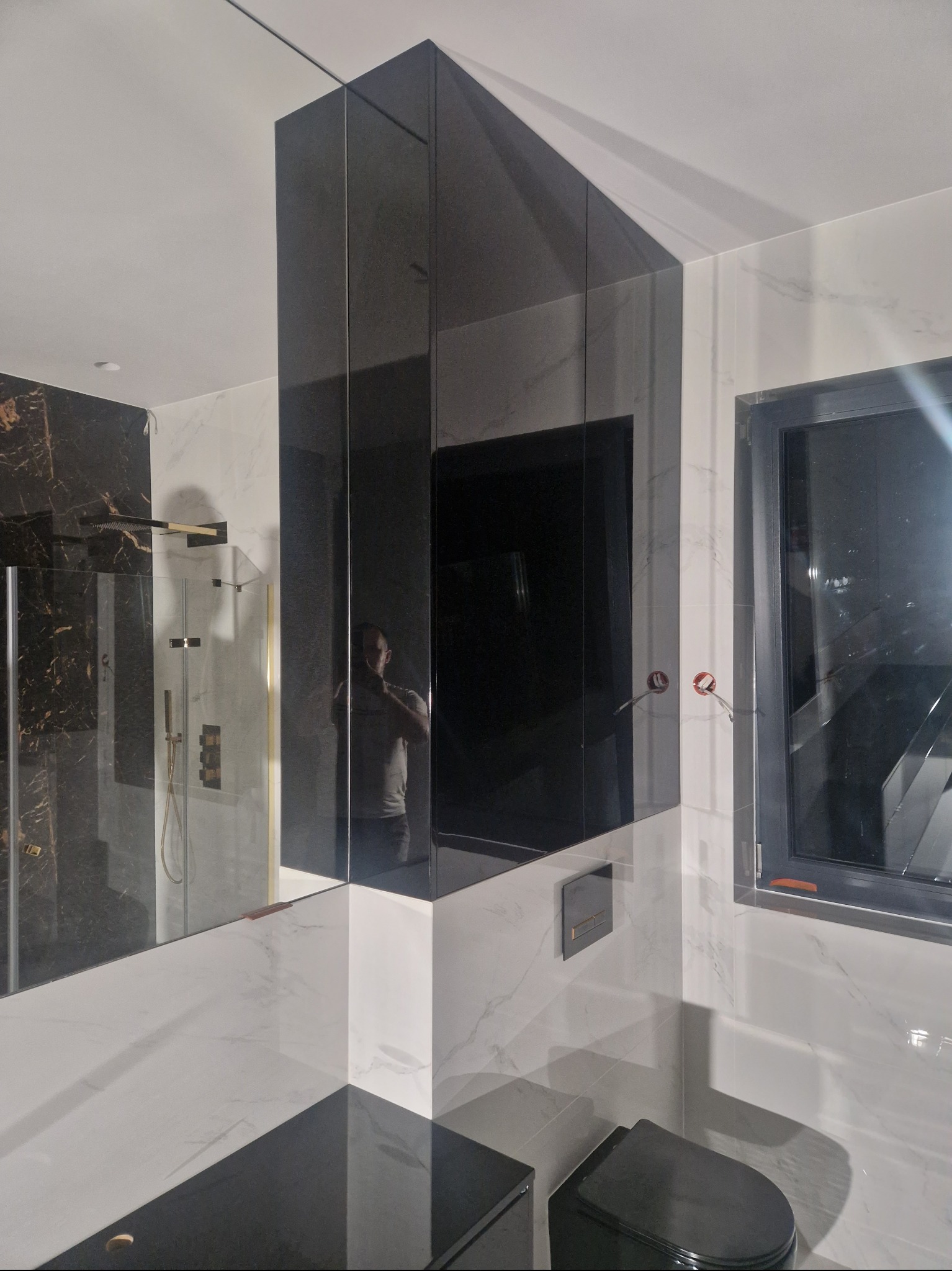 Nowoczesna łazienka z czarnymi, lustrzanymi szafkami narożnymi, białymi marmurowymi płytkami ściennymi i czarną armaturą, w tym toaletą i oknem.