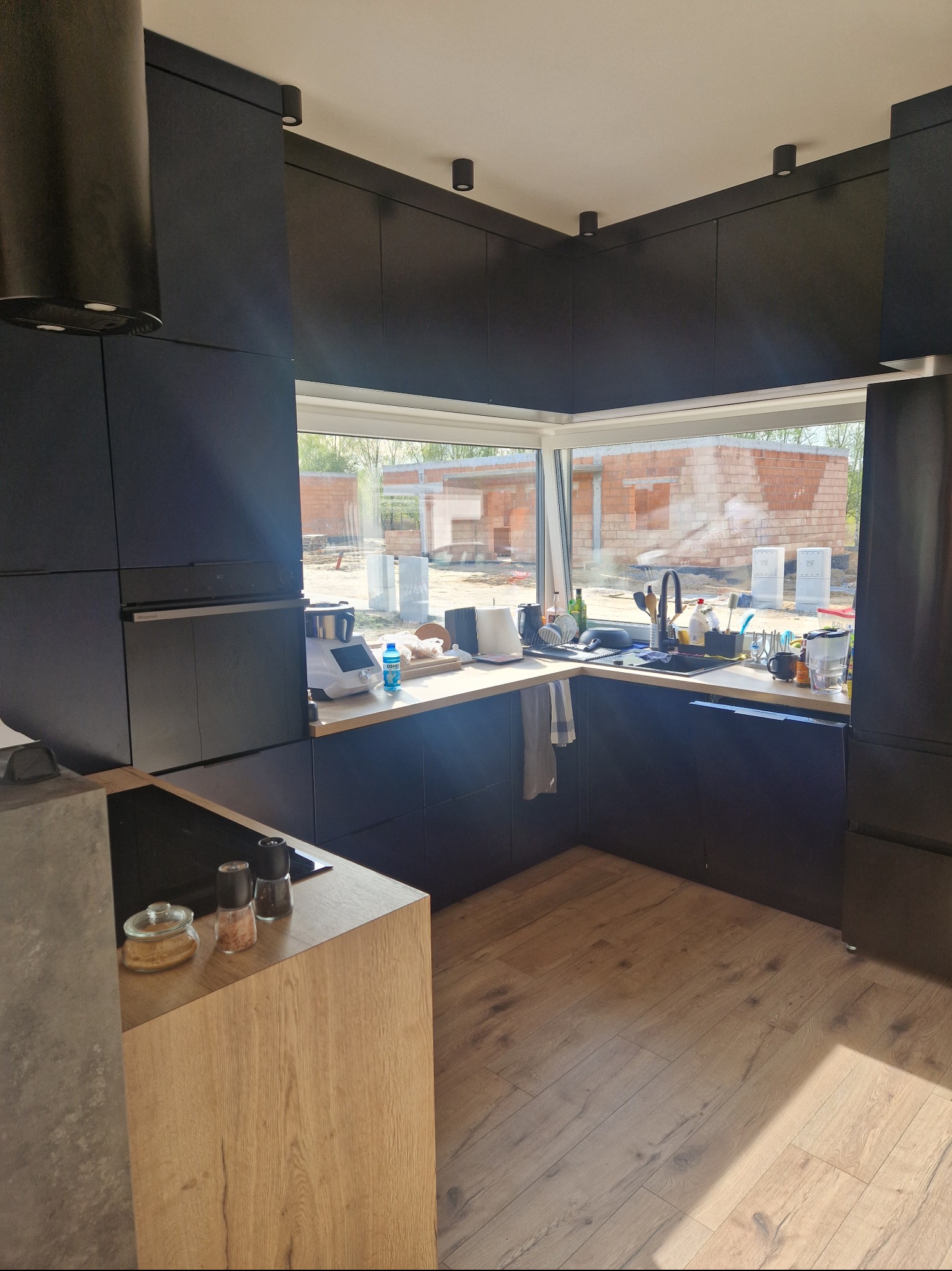 Nowoczesna, czarna kuchnia z drewnianymi blatami w Aleksandrowie Łódzkim, widok na budowę przez duże okno, minimalistyczny design.