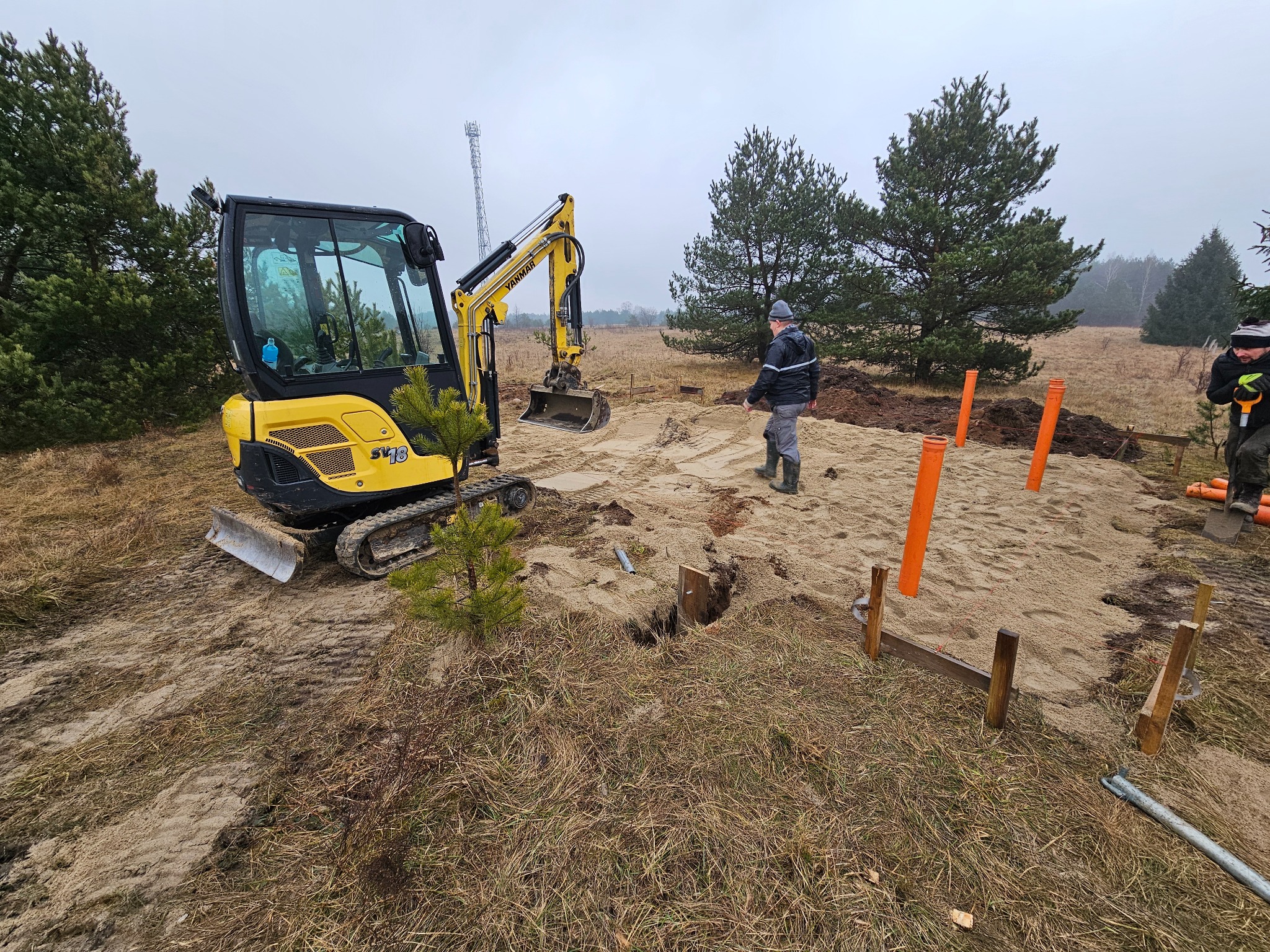 Żółta mini koparka Yanmar SV18 na piaszczystym terenie budowy z wyznaczonym obszarem, widoczne pomarańczowe rury i drewniane szalunki, dwóch pracowników w oddali, pochmurne niebo.