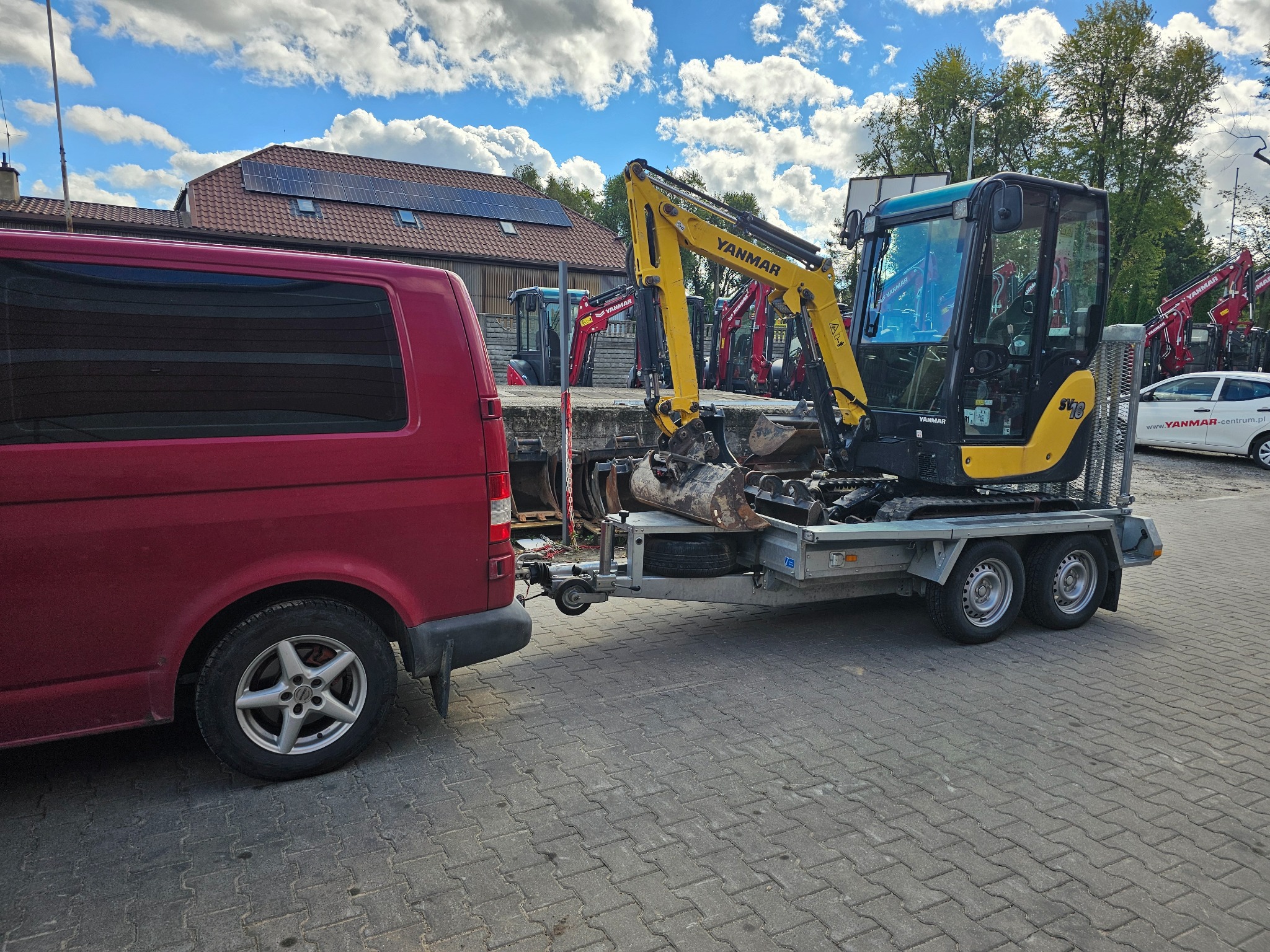 Czerwony bus holuje na przyczepie małą żółto-czarną koparkę Yanmar SV18 na tle innych koparek i budynku z panelami słonecznymi.