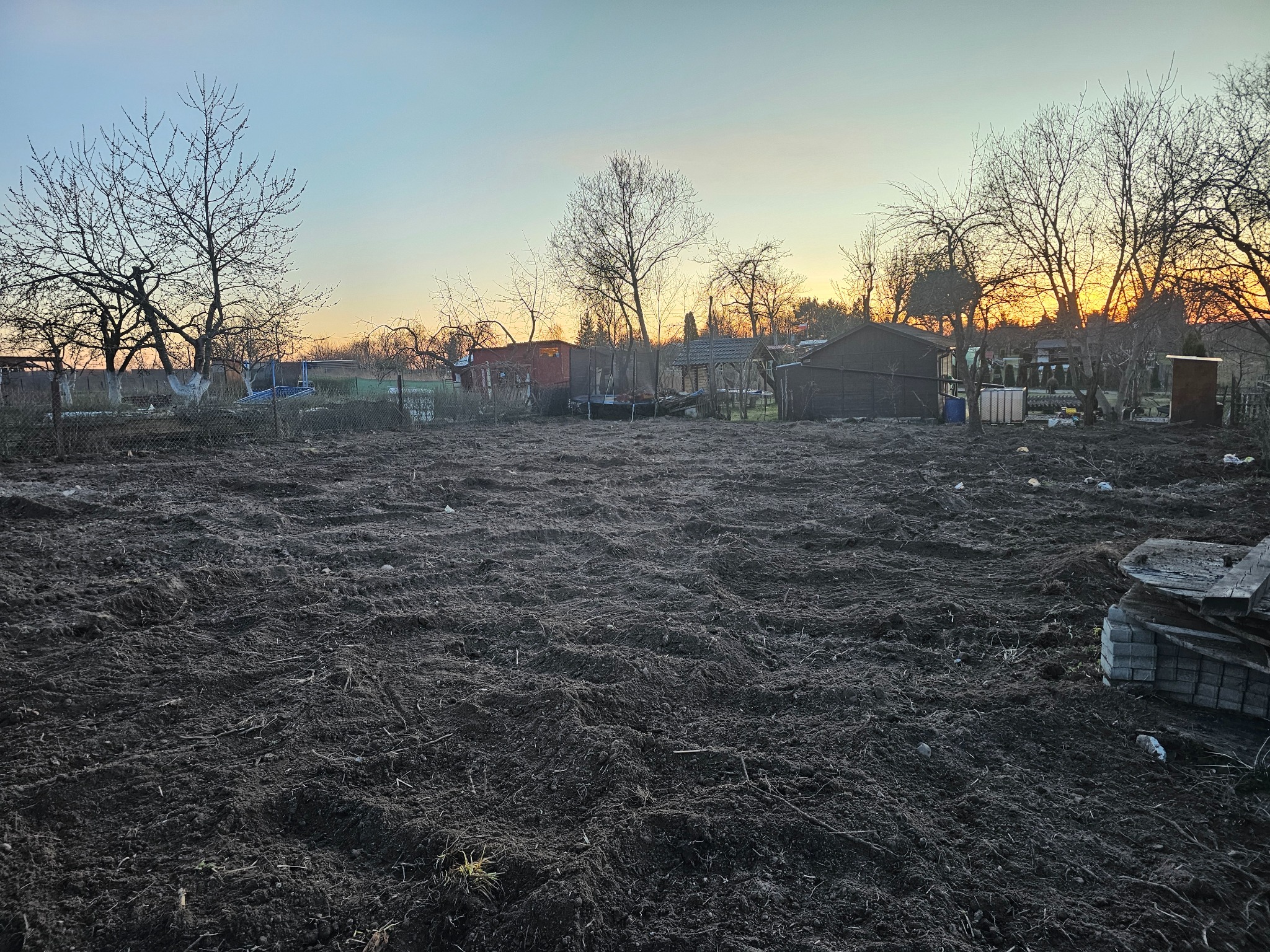 Przygotowany teren pod budowę z widocznymi śladami po maszynach, w tle zabudowania gospodarcze i drzewa o zachodzie słońca.