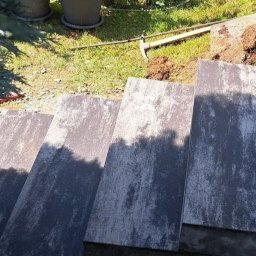 ZABRUKOWANI Dominik Dubiel - Schody zewnętrzne wyłożone szarą kostką granitową, widok z góry z fragmentem iglastego krzewu po lewej stronie i narzędziem ogrodniczym oraz ziemią w tle.