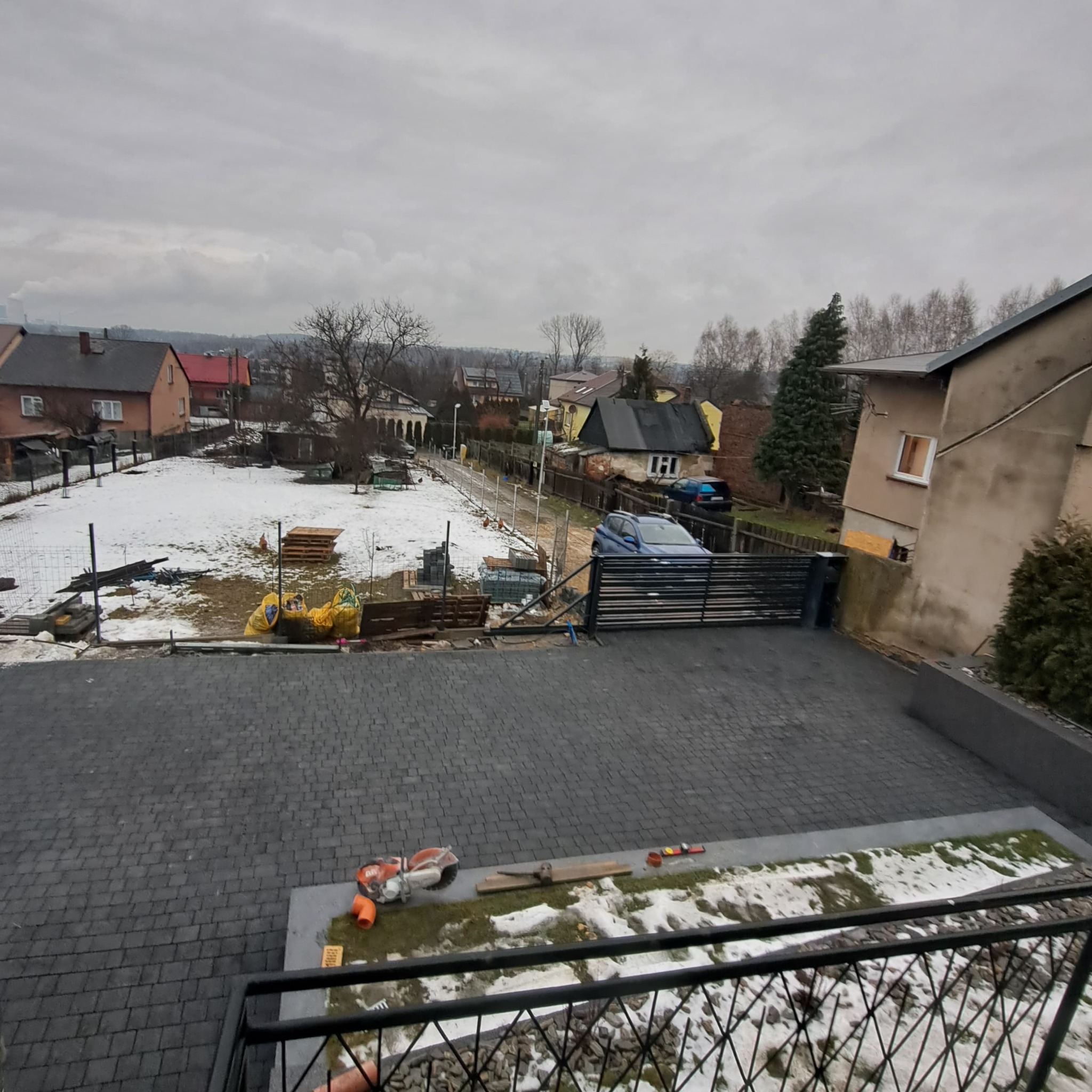 Widok z góry na posesję w trakcie prac brukarskich, z widoczną ułożoną kostką, narzędziami, ogrodzeniem i placem z przygotowaniami pod dalsze prace, wszystko w zimowej scenerii.