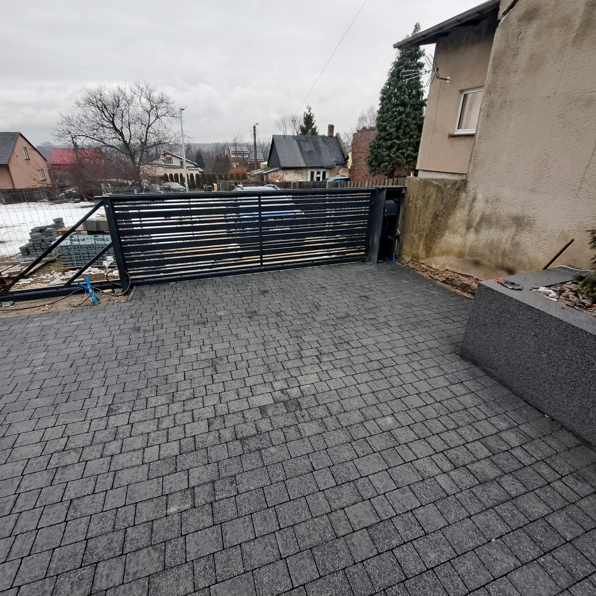 Ułożona, szara kostka brukowa na podjeździe z czarną, przesuwną bramą w tle, widoczne przygotowane kostki do dalszej pracy.