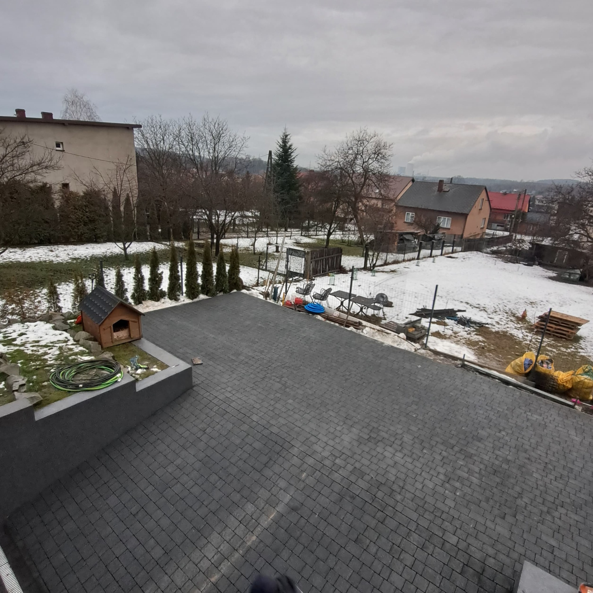 Ułożona szara kostka brukowa na posesji w pochmurny dzień, widok z góry, w tle domy i resztki śniegu, obok kostki psia buda i zwinięty wąż ogrodowy.