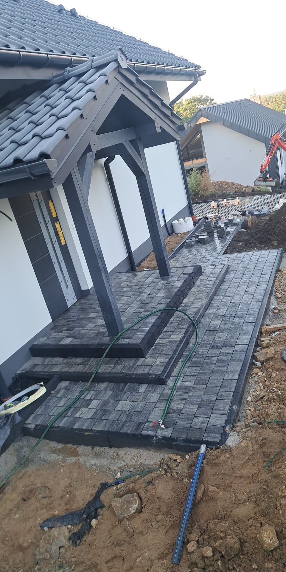 Układanie szarej kostki granitowej przy wejściu do domu z ciemnymi drewnianymi słupami, widoczny fragment ogrodu i koparka w tle.