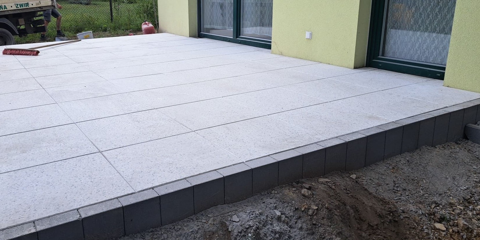 Nowo ułożony, jasnoszary taras z płyt betonowych, obrzeża z ciemnych kostek, częściowo widoczny budynek z oknami i zielonkawą elewacją, ziemia wokół tarasu jeszcze niezagospodarowana.